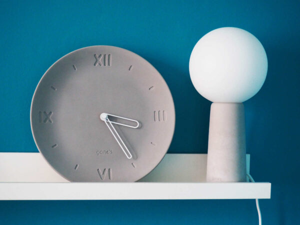 Horloge forme assiette béton gris clair chiffre romain aiguilles blanches côté lampe béton ampoule blanche