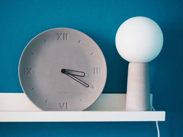 Horloge forme assiette béton gris clair chiffre romain aiguilles noires côté lampe béton ampoule blanche