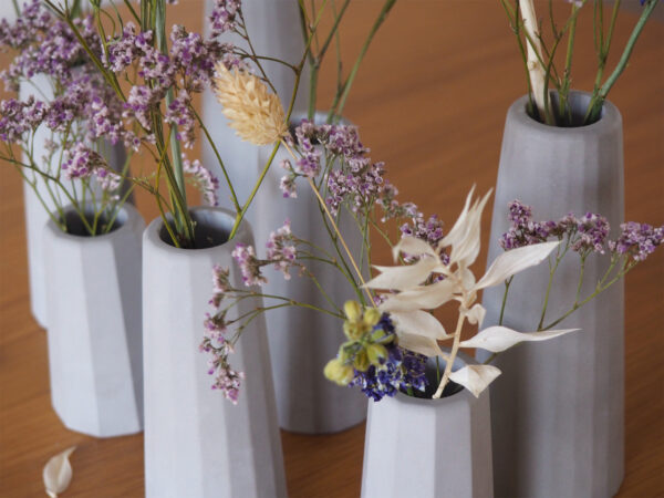 Plan sept petits vases béton gris clair différentes tailles surfaces facettes composition milieu table bois fleurs