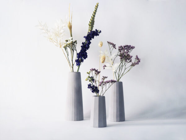 Trois petits vases béton gris clair différentes tailles surfaces facettes contentant fleurs séchées.